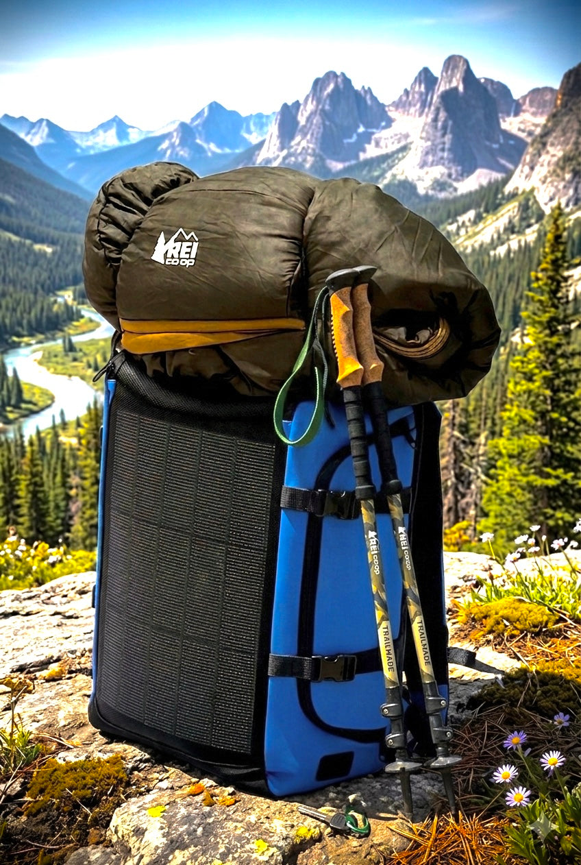 40Fied blue Fortified Solar Everyday Backpack with integrated 15 Watt solar panel, staged on a Rocky Mountain overlook with sleeping bag and trekking poles, ideal for hiking, camping, and off-grid adventure